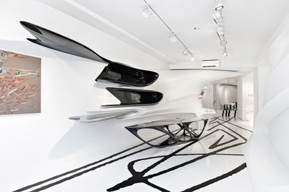 Galerie Gmurzynska Gallery. The room is all white, with black lines on the floor. A metallic, modern table, asymmetrical in shape with matching shelves on the wall. There is an abstract painting to the left.