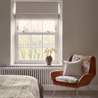 White bedroom facing the wooden windows. Next to the window is an orange accent chair