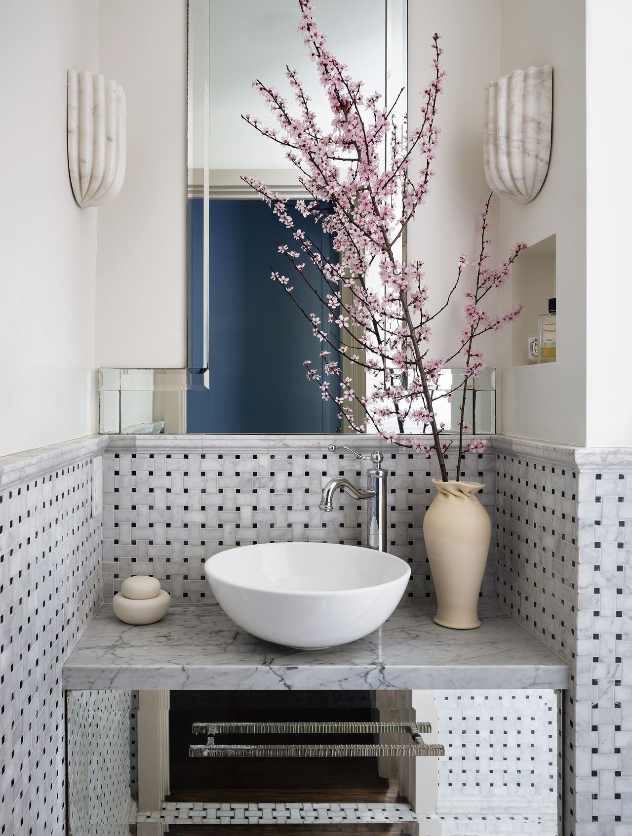 Image of a bathroom vanity area where half of the wall is tiled in a basket weave style. There is a mirror on the wall and two marble sconce lights. There is a vase on the countertop with a large pink flowered stem.