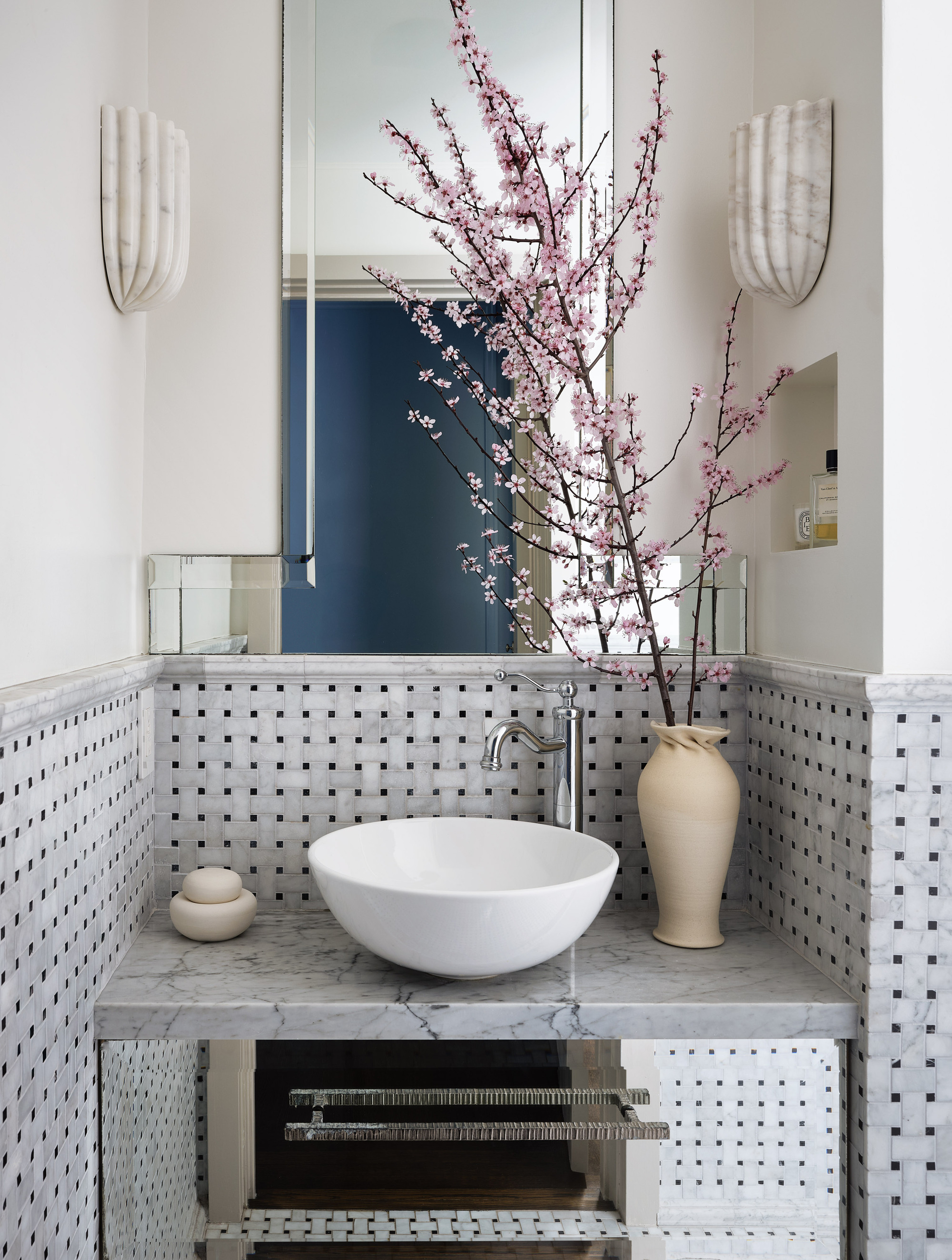 Image of a bathroom vanity area where half of the wall is tiled in a basket weave style. There is a mirror on the wall and two marble sconce lights. There is a vase on the countertop with a large pink flowered stem.