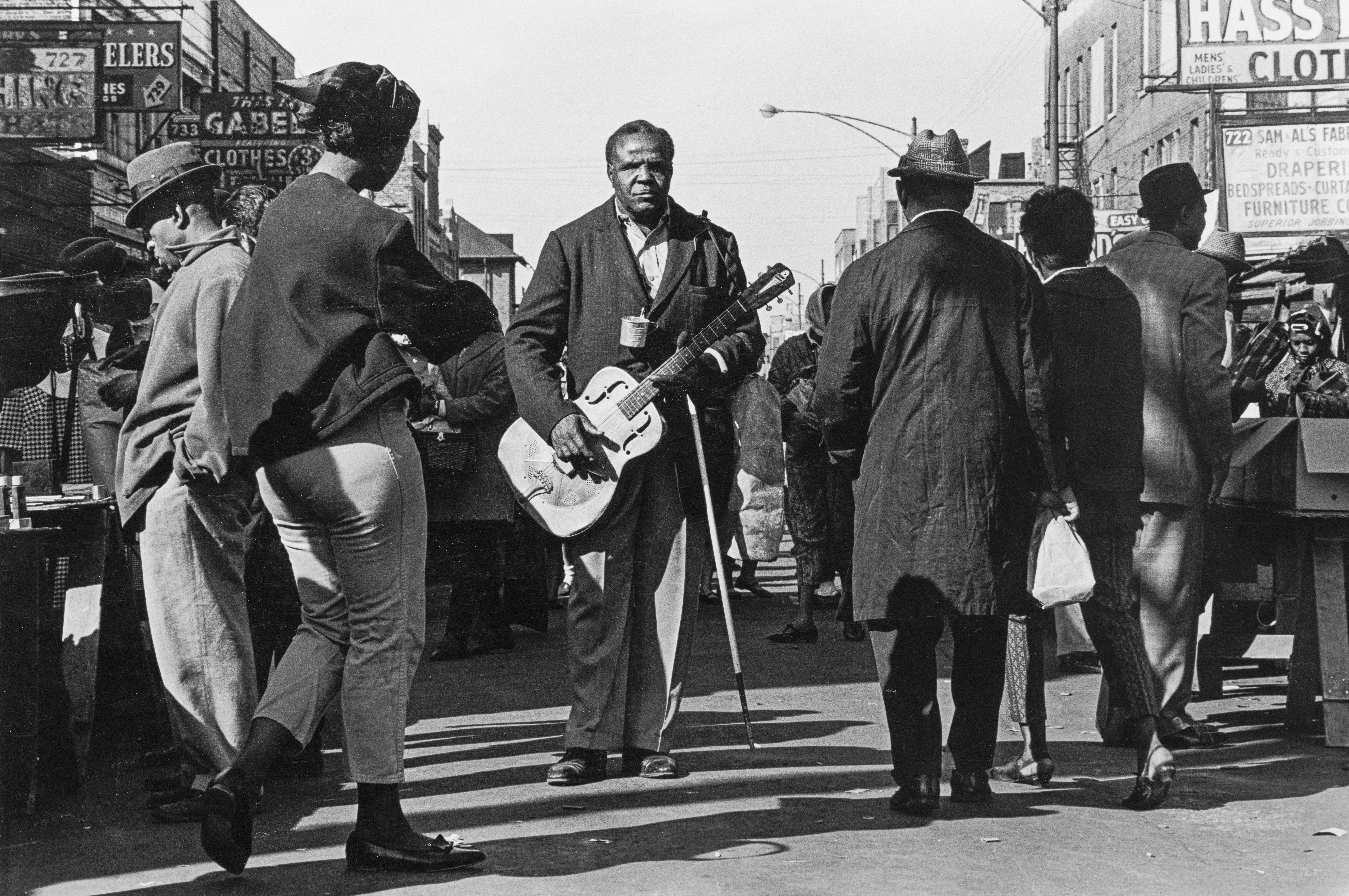 A black and white street photograph of a man standing in the middle of a crowded Maxwell Street Market holding a resonator guitar.