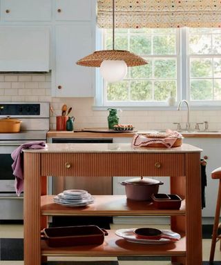 A kitchen with a wooden rectangular kitchen island with a rattan pendant light above it and light blue cabinets and a window with beige blinds behind it