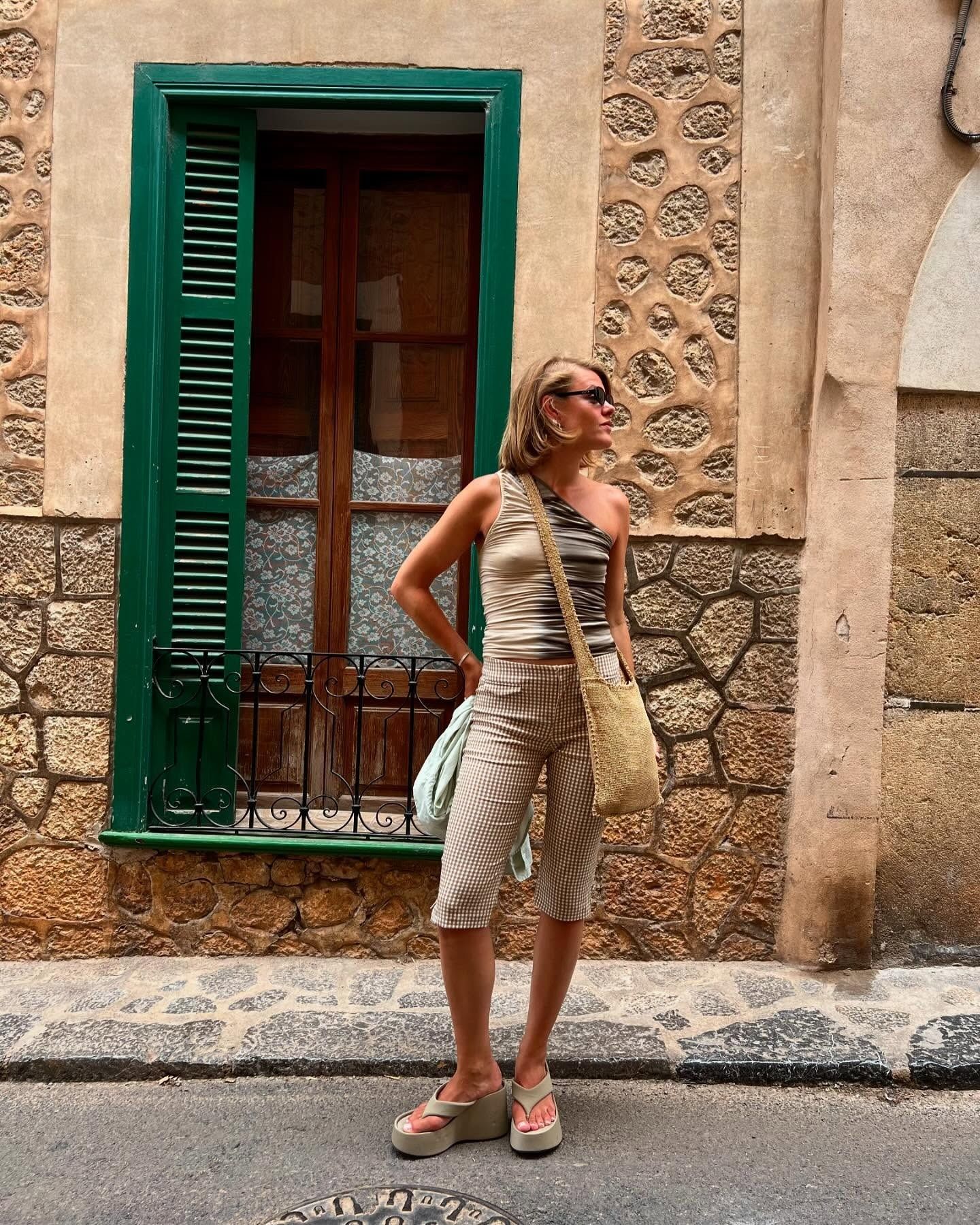 Influencer wears platform flip-flops with capri pants and a one-shoulder top. She's styling a raffia cross-body bag over her shoulders and is wearing dark sunglasses.