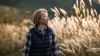 A woman in a navy blue gilet and plaid shirt walks through a field of tall plants. The sunlight hits her face, which is slightly turned away from camera and she is smiling slightly. Her hair is bobbed just above the shoulder and is a light copper color.