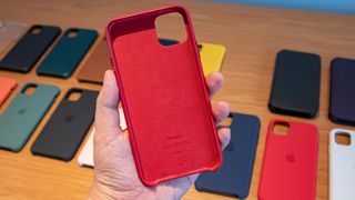 En hand håller upp ett rött skal till en iPhone 11, med ett gäng andra skal i olika färger uppradade på ett bord i bakgrunden.
