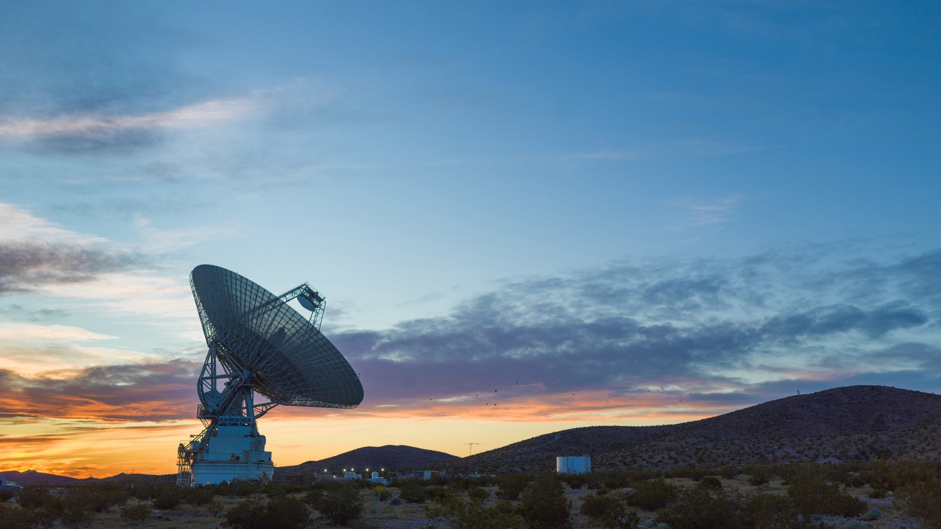 a large radar antenna dish in the desert points upwards towards the sky at sunset