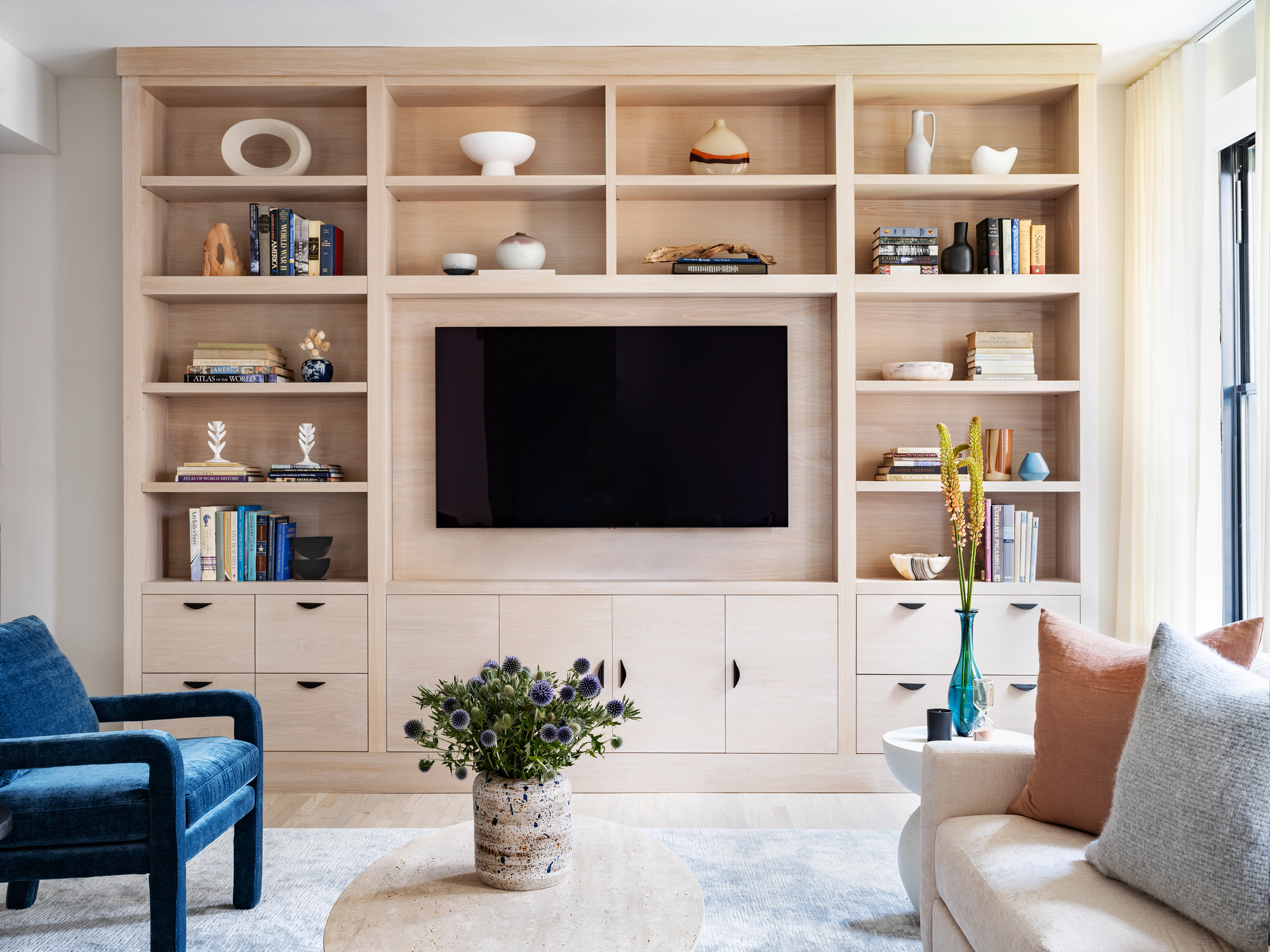 modern living room with built-in wall shelving, mounted tv, blue armchair opposite a white sofa with pillows, and a travertine coffee table with vase of flowers