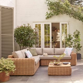 A wicker-effect outdoor sofa with cream cushions on a paved patio area in front of a house