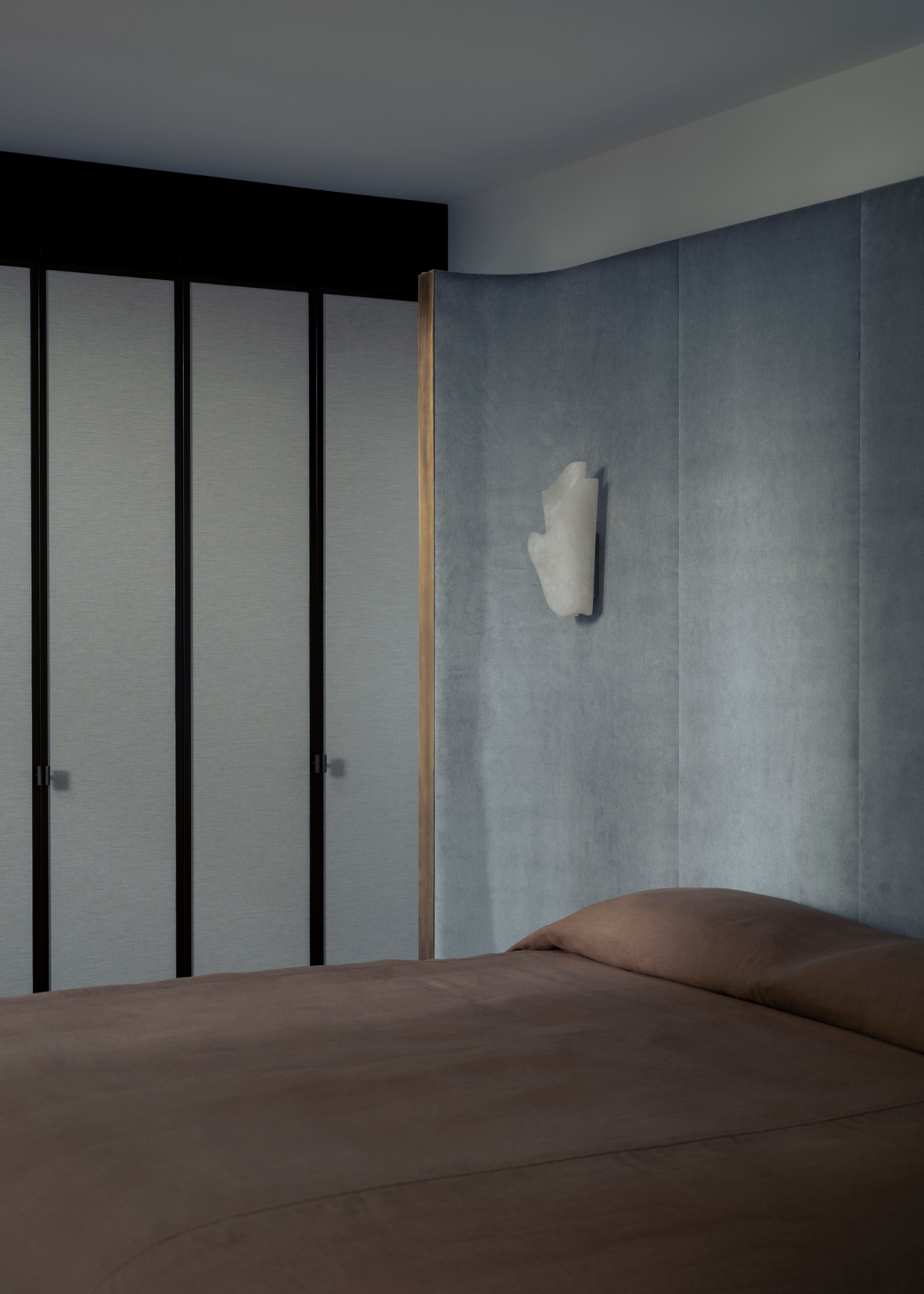 A dimly lit bedroom with an oversized dusty blue headboard, a wall-mounted sconce and brown sheets