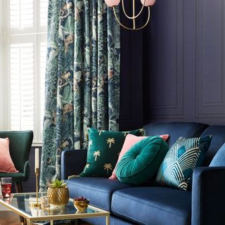 living room with dark blue walls and rich blue sofa with green, blue and red cushions, a gold glass top coffee table and jungle green patterned curtains