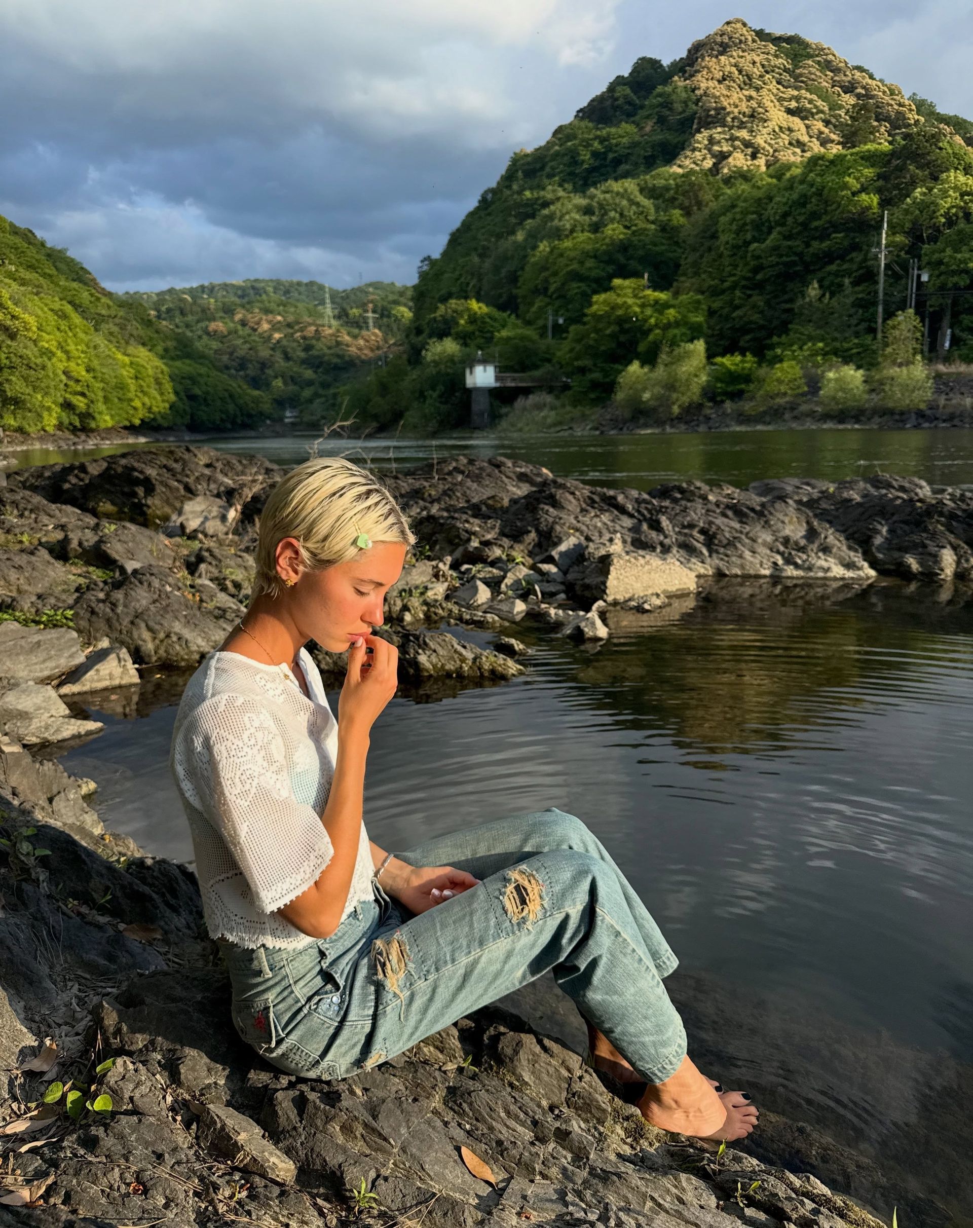 Iris Law sits by a lake wearing ripped jeans with a white boho blouse.