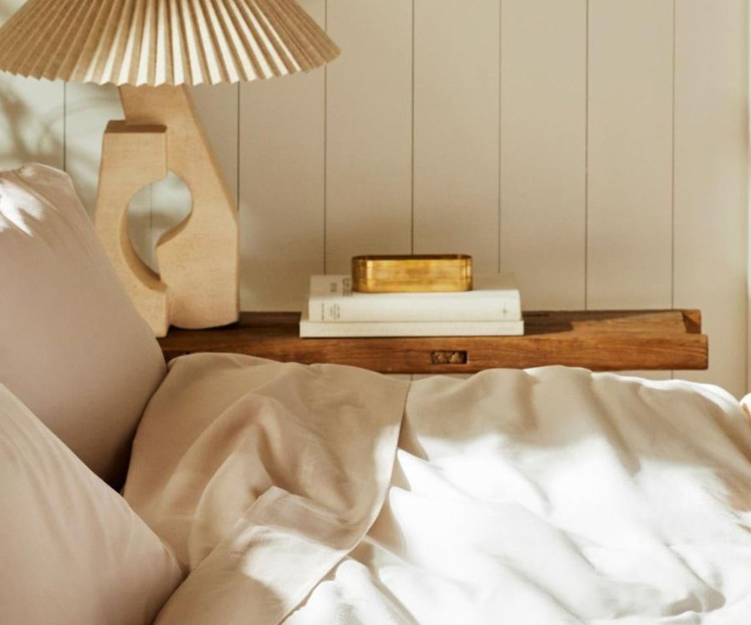 Brooklinen sateen sheets on a bed; in the background, a lamp and books on a nightstand against cream panelled walls.