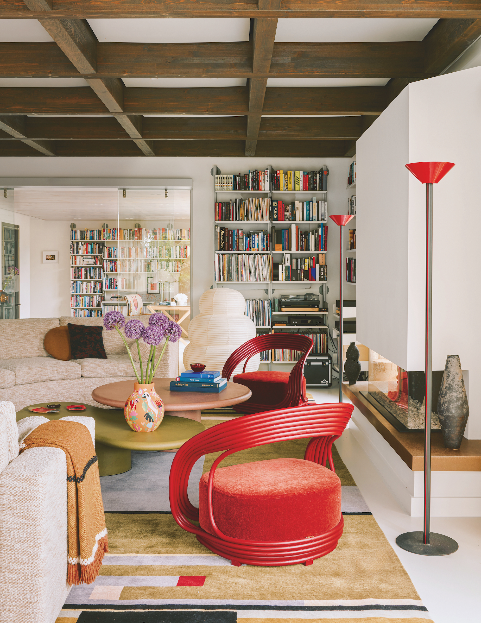 A modern, eclectic style living room with exposed beamed ceilings. white walls, and large book cases on the two far walls. The seating area is comprised of one large curved sofa, two red armchairs and a multi-level coffee table.