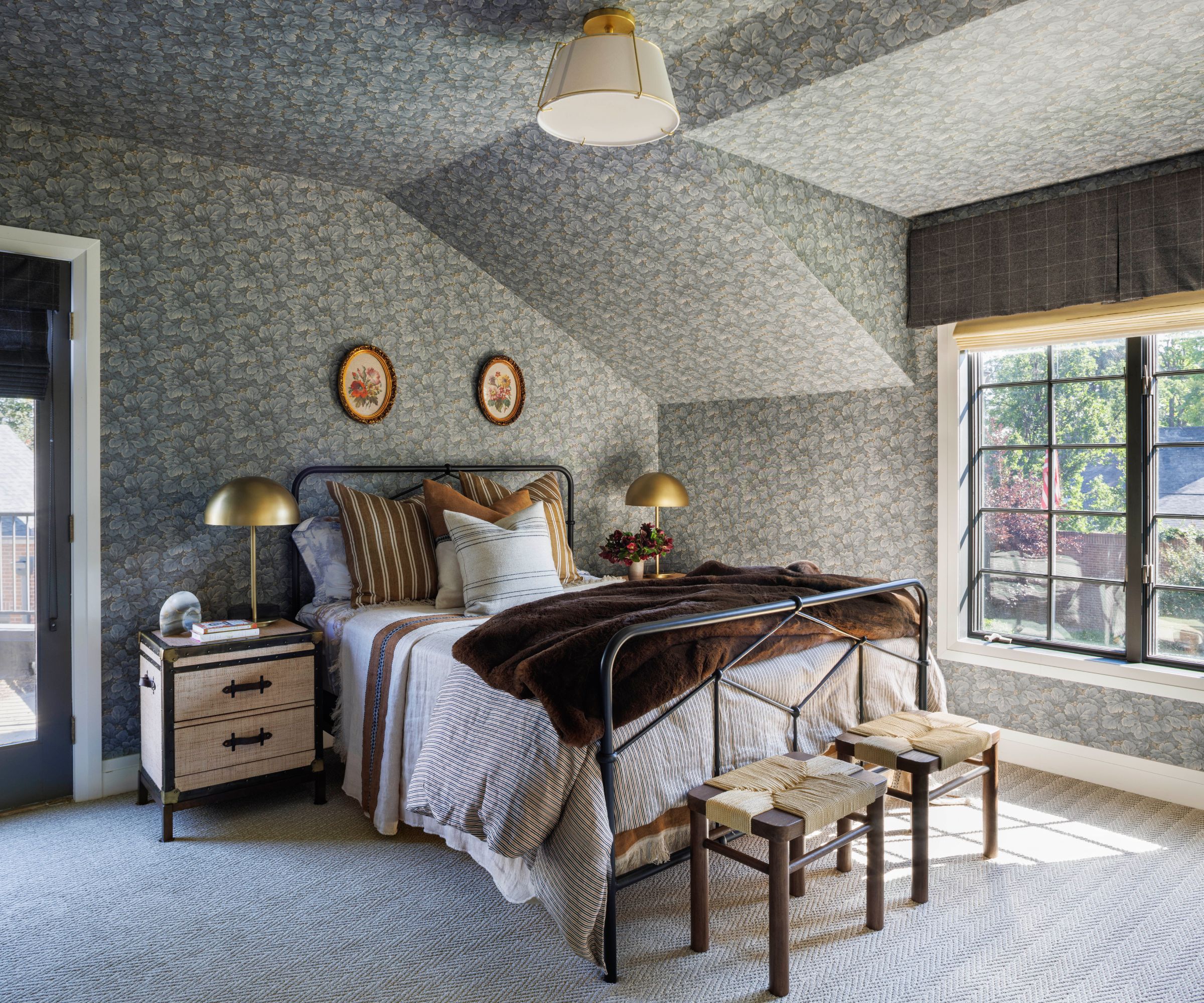 Bedroom with gray floral wallpaper over walls and ceiling with black iron bed frame and two wood and rattan stools at the bottom of the bed