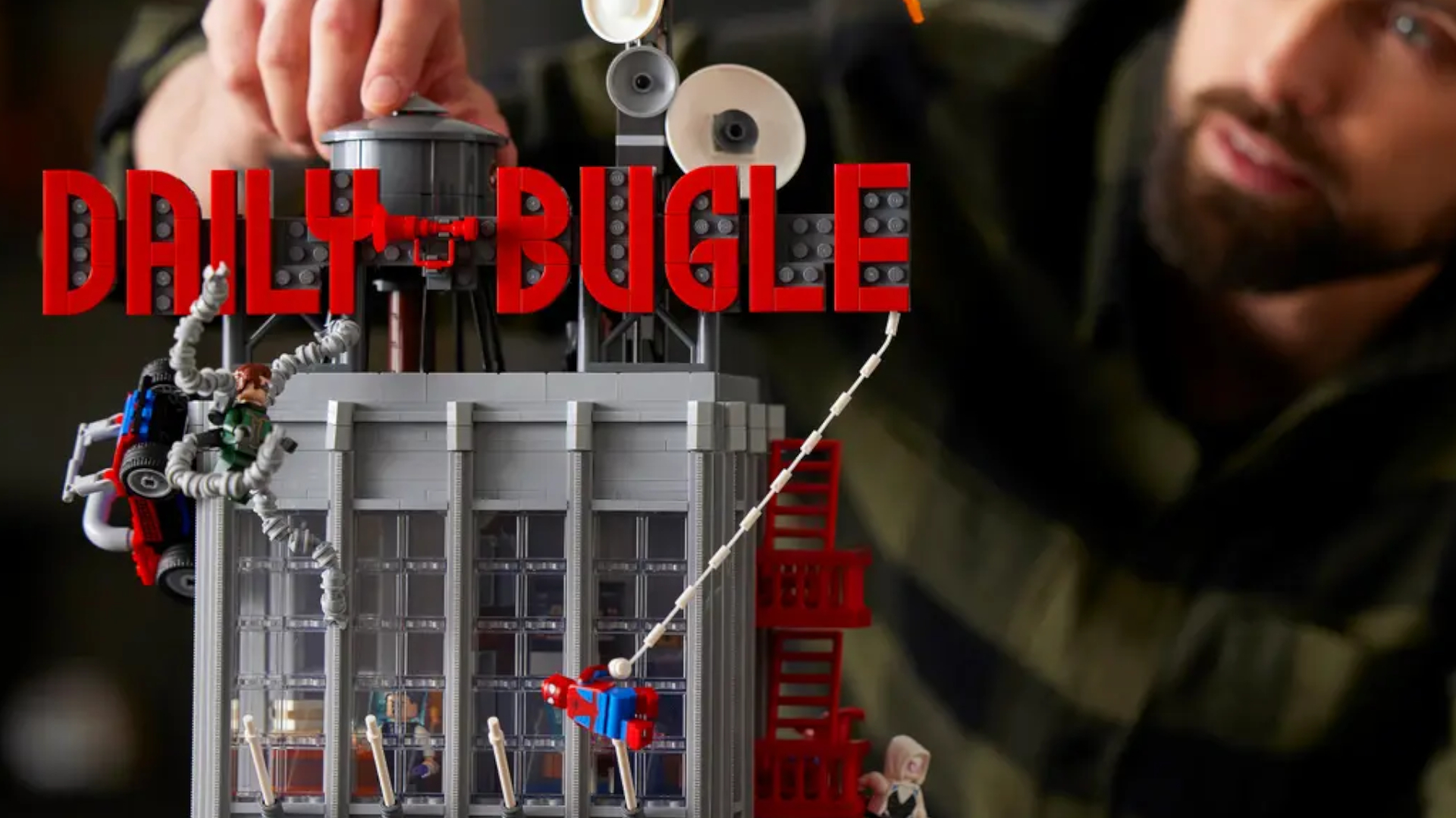 A bearded man smiles as he places a piece onto the Lego Daily Bugle set, which has minifigures swinging from it