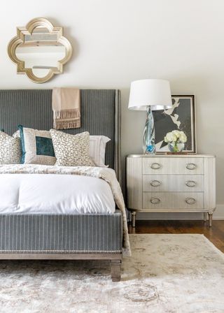 white bedroom with grey upholstered stripe bed, with bedding, rug, wooden floor, vintage bedside