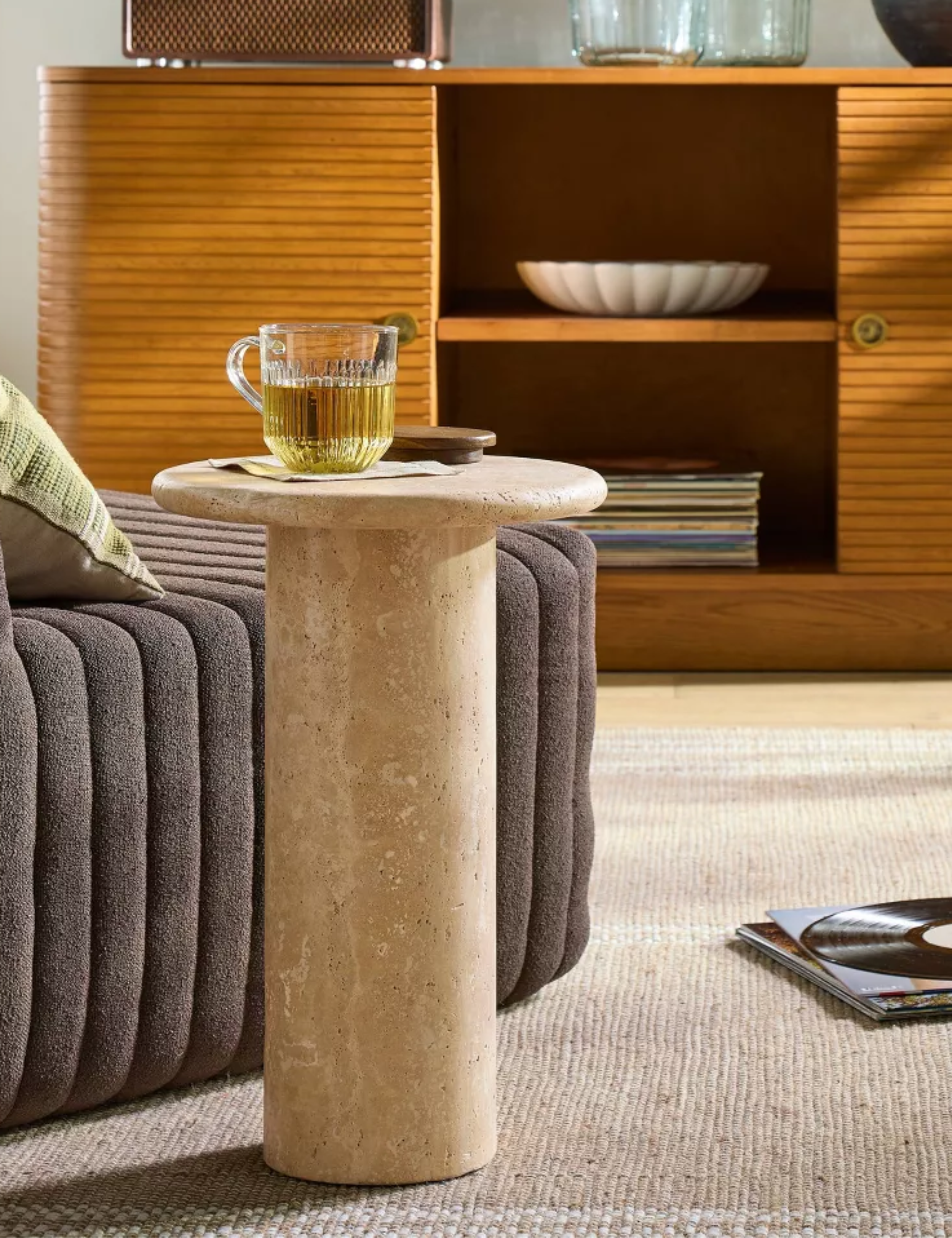 Joanna Gaines travertine table sitting alongside a channel tufted accent chair in a carpeted living room that features a fluted sideboard