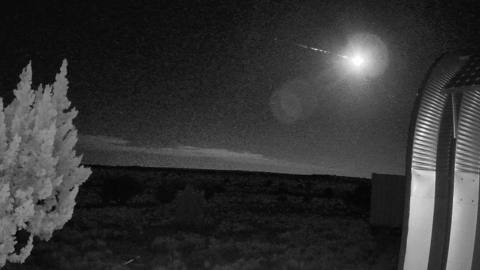 A fireball is pictured streaking through the night sky above a field. A steel building is visible to the right of the image, and a bush frames the left of the shot.