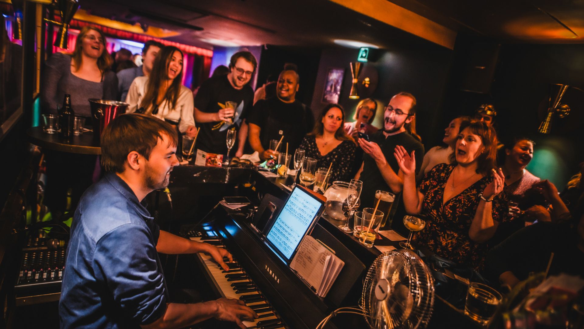 an image of a pianist playing the piano with a group of people singing around it in a bar