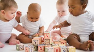 Four babies sitting on floor playing with colorful number and letter blocks.