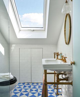 Bright white bathroom with white painted walls and ceiling, blue and white geometric floor tiles, skylight on slanted ceiling, traditional white sink with exposed metal piping, metal rounded mirror and metal and glass wall light