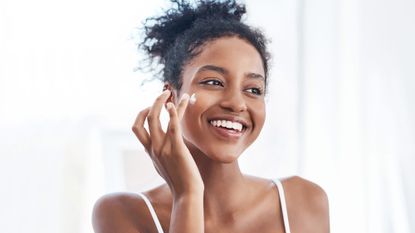 An image of a woman applying a skincare product on her cheek whilst smiling