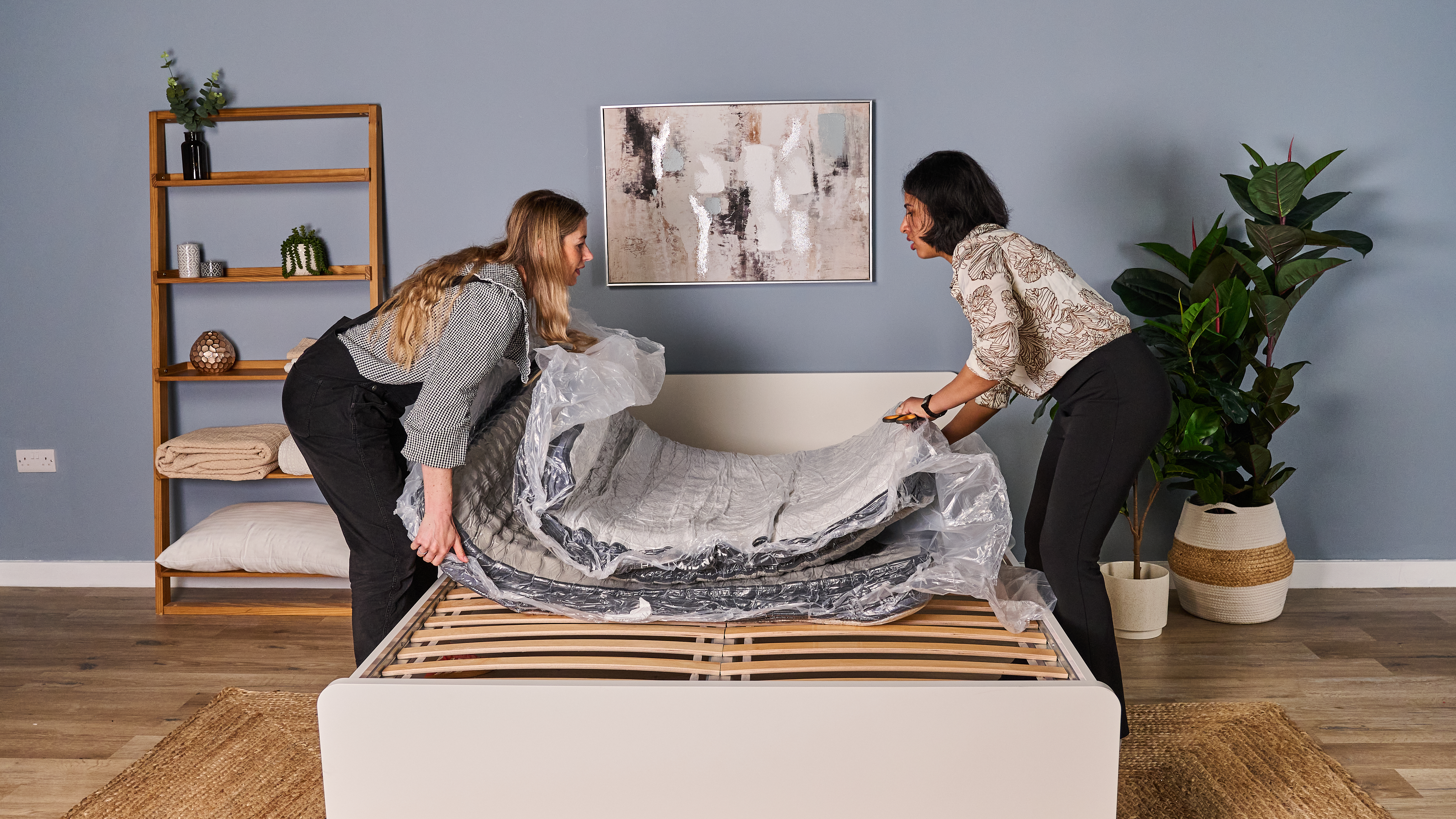 The image shows two mattress testers unboxing the Helix Midnight Luxe mattress and setting it up on a white bed frame in a blue bedroom