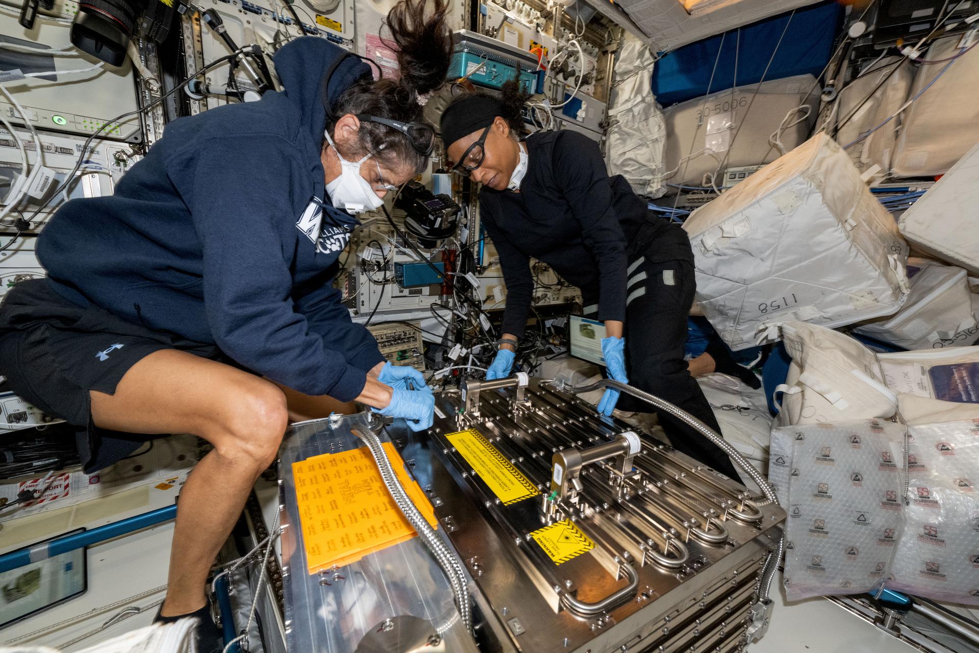 Dos mujeres en chándal desempacan una gran caja de metal en un laboratorio estrecho lleno de cables y computadoras. ellos flotan