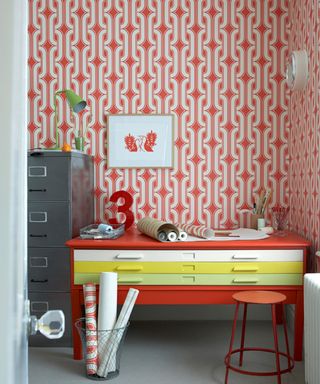 bold red wallpaper in home office, and a multi-colored desk with drawers, a red stool, and a bin full of wrapping paper and a gray filing cabinet to the right