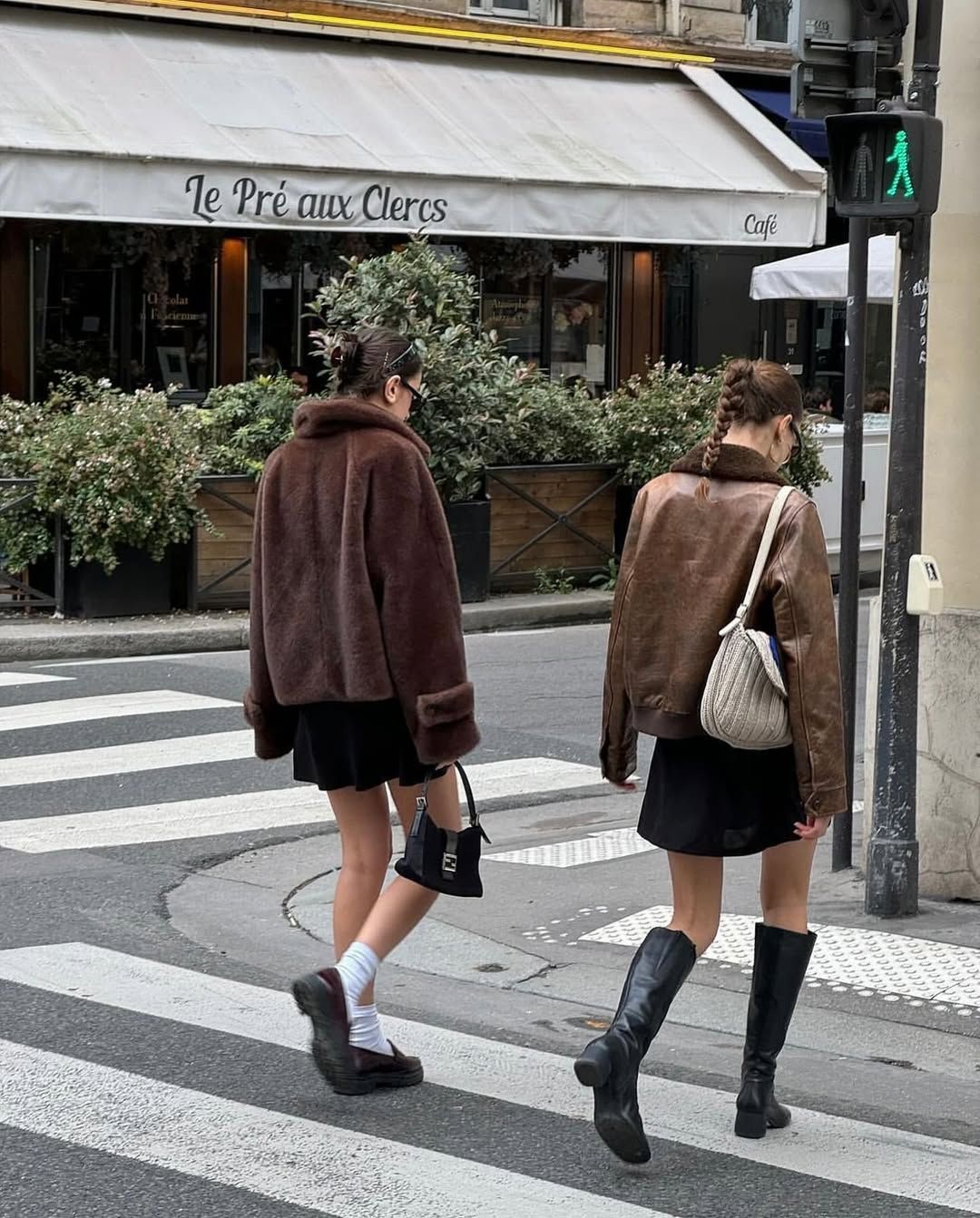 Two women crossing street in brown jackets and black skirts.