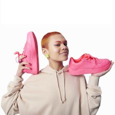 a woman with short orange and pink hair in a beige hoodie, holding a pair of pink trainers in her hands