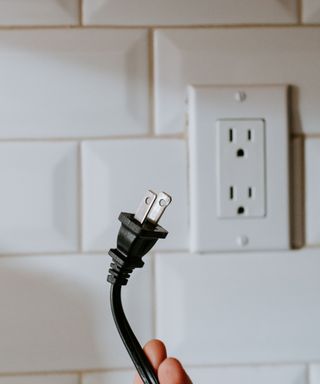 A black, american plug being held in front of a white outlet in a white tiled wall