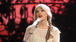 Taylor swift, wearing her hair in pigtails with a cream colored sweater, standing at a microphone in front of a black and red background