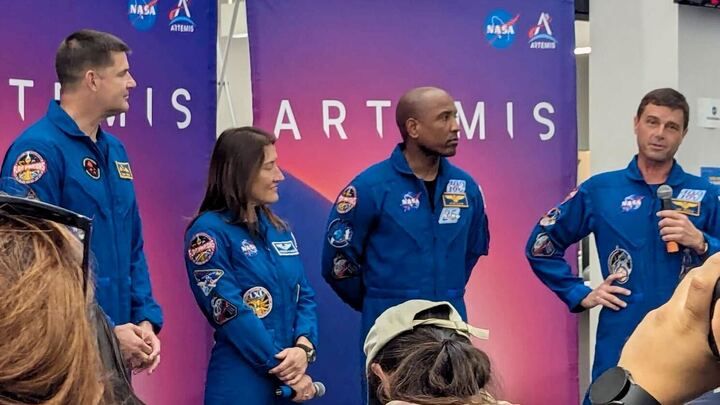 four people in blue flight suits stand in a room. One holds a microphone.
