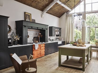 large kitchen with orange range, sage green kitchen island, open door to outside, clear glass pendant lights, charcoal cabinetry, leather armchair, drapes