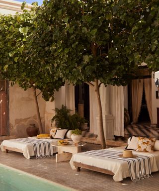 A backyard area with two white sunloungers on it, a white marble table between them, two tall trees above them, a white building behind them, and a swimming pool in front