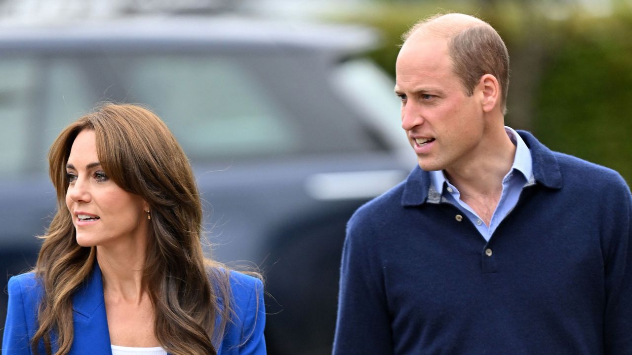 Kate Middleton wearing a blue blazer and walking in front of a car next to Prince William, who is wearing a blue sweater, with both looking to the left 