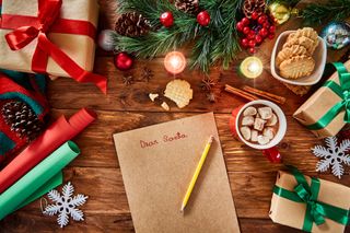 An image taken from above of a sheet of brown paper with the words 'Dear Santa' written in red, surrounded by Christmas decorations such as presents, cinnamon sticks, a hot chocolate, and some fresh pine.