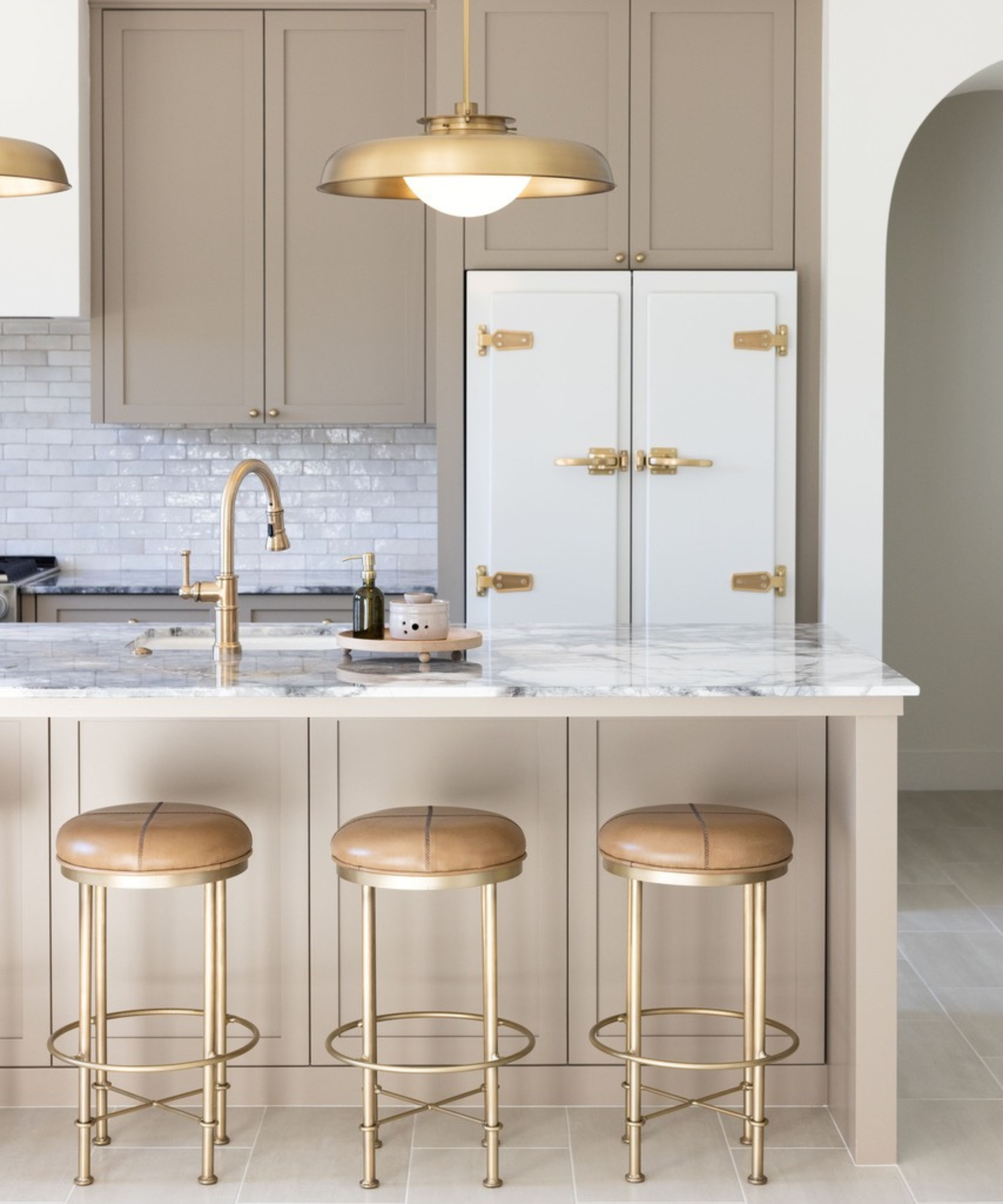 A sophisticated kitchen island with taupe cabinetry and three leather-topped brass barstools, featuring a white retro-style refrigerator with gold hardware in the background