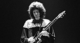 A black-and-white photo of Brian May of Queen playing his Red Special onstage in 1974 as the band tour their sophomore album Queen II. 