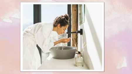 An image of a woman in a robe washing her face by a bathroom sink on a pink marbled background