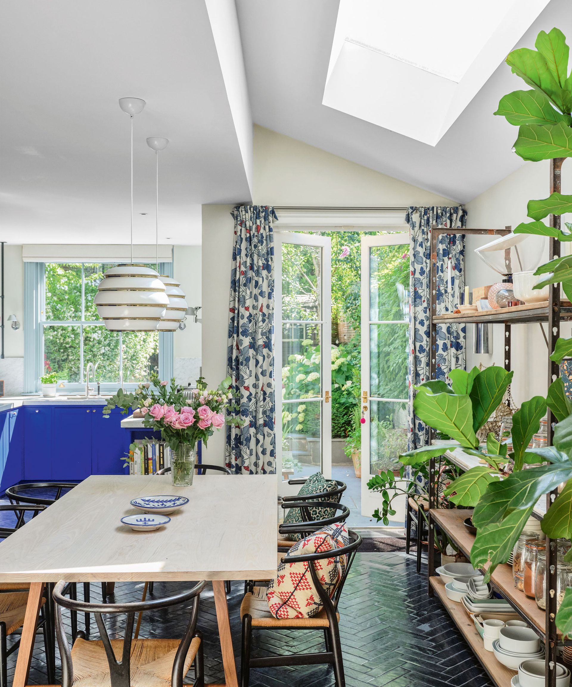 Open plan kitchen dining room, black herringbone tiled flooring, white painted walls, skylight on ceiling, lightwood dining table with black wood dining chairs, floral curtains, large shelving unit decorated with kitchenware, two white hanging pendants above dining table