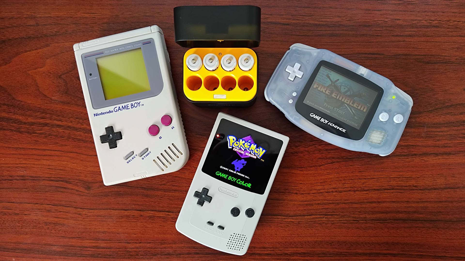 Rechargeable AA batteries in black and yellow case with Game Boy on left, modded Game Boy Color below, and Game Boy Advance on right sitting on woodgrain desk.