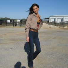 Bella Hadid wearing an open button down top with blue jeans a brown belt and western boots