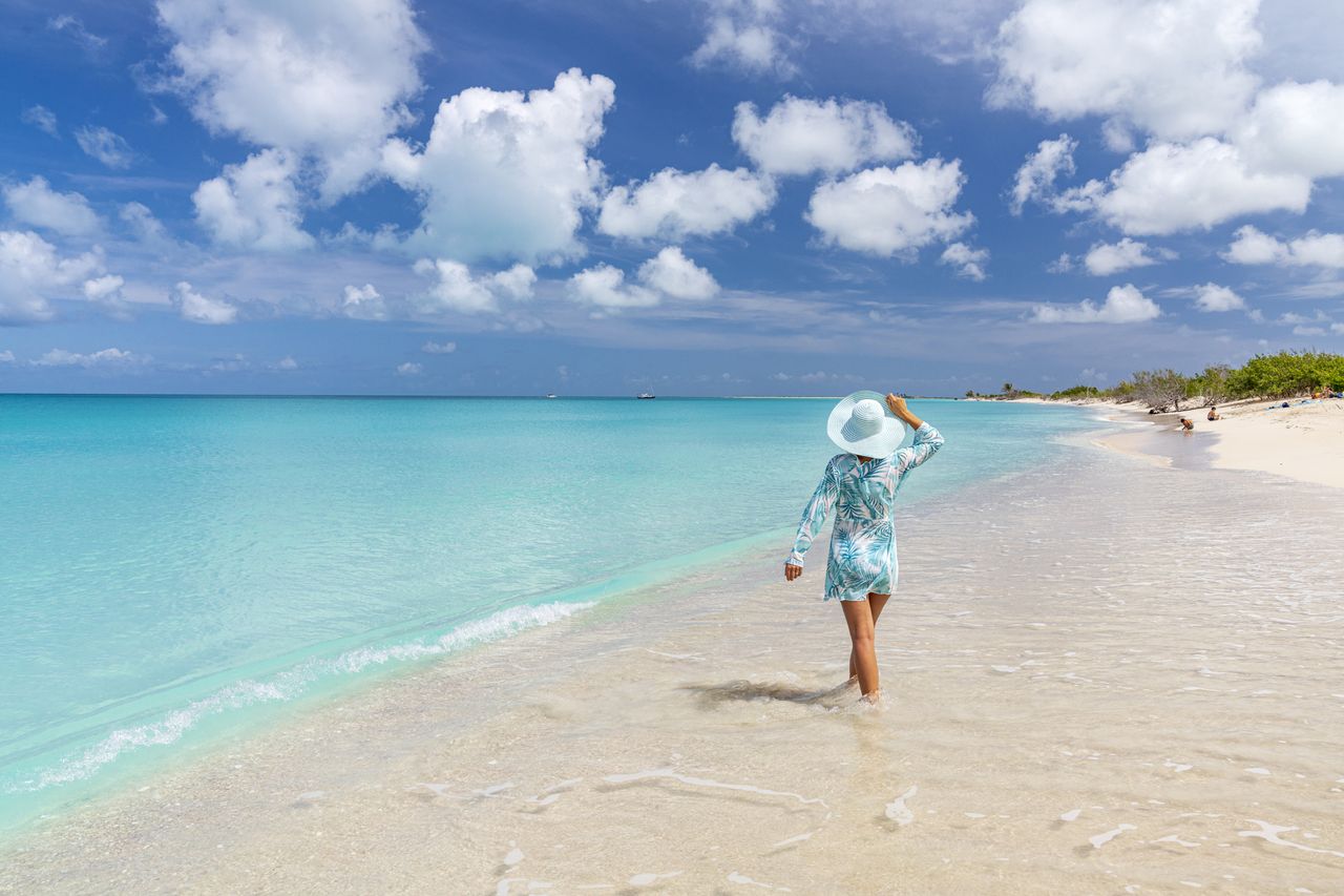 An older woman walks away from the camera on a tropical beach as part of post-holiday travel.