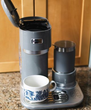 Mr. Coffee 4-in-1 Single-Serve Latte, Iced, and Hot Coffee Maker in grey colorway on granite-effect worktop with blue and white chinoiserie-effect ceramic mug
