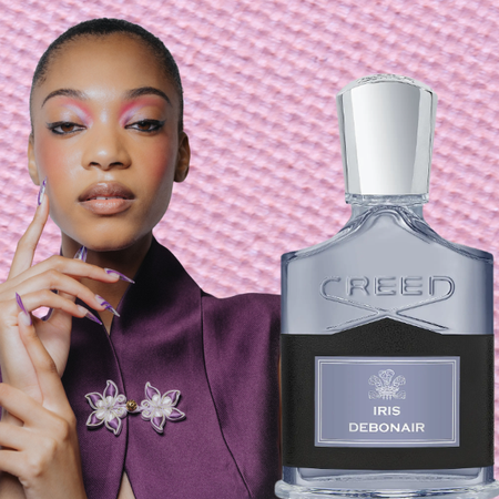 A woman wearing a purple tops, nails, and makeup next two two bottles of perfume all in front of a textured, light purple eyeshadow background. 