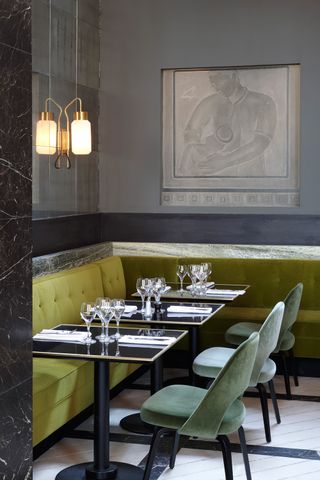 A sleekly furnished, modern restaurant features soft, plush seating in different shades of green, grey walls adorned with artworks, and laminated glass lighting.