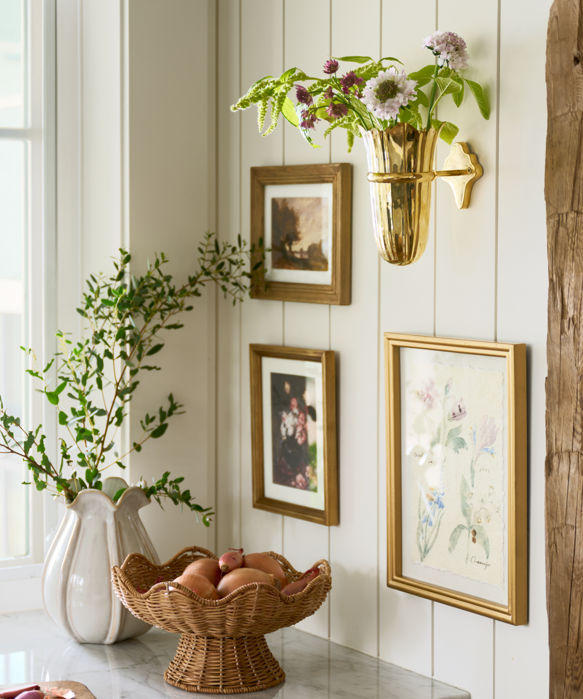 a countertop corner with photos hanging, a wall-mounted vase, a vase with branches, and a rattan fruit bowl