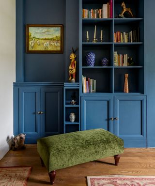 dark blue painted storage and display unit, built in shelving with books and ornaments, dark wooden flooring with two rugs, green fabric ottoman with dark wood legs